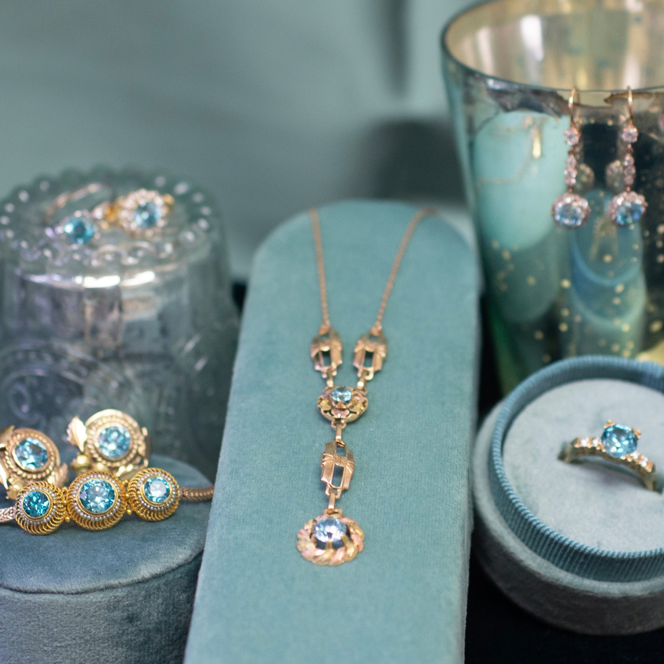 Blue zircon and yellow gold including several rings, a necklace, bracelets and earrings displayed on a soft teal surface with a blurred background. All pieces are curated by The Jeweled Crescent in Portland, Oregon
